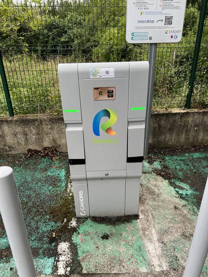 Electric vehicle charging station by Révéo with green indicator lights, situated on a weathered concrete surface, next to an informational sign.