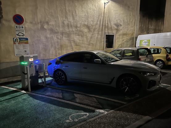 A BMW i4 electric car charging at a public charging station at night, with nearby parked cars and a wall with signs.