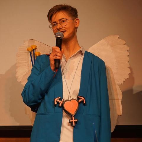 One person on stage with a mic in the hand and wearing a blue blazer and wings with bow and arrow. Around the neck they are wearing a necklace with a heart and "all gender" arrows.