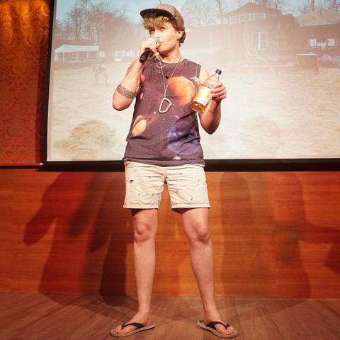 Person with mic on stage. Wearing a carabiner on a necklace, holding a bottle in their hand.