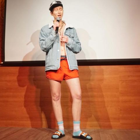 One person standing on stage with a mic in the hand and wearing a cap, sports pants and a jeans jacket.