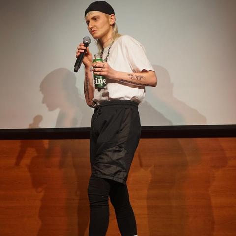 One person in stage with a beanie and a white shirt holding a beer in one hand and a mic in the other.