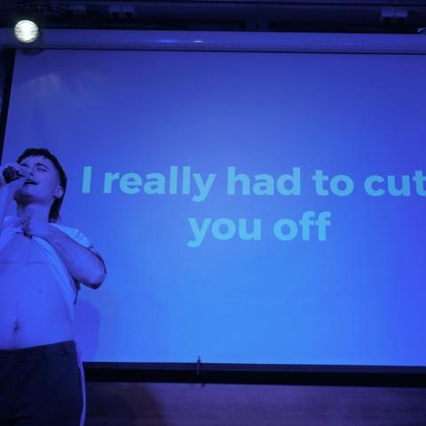 Person standing on stage, singing into the mic and lifting their shirt. The background screen reads "I really had to cut you off".