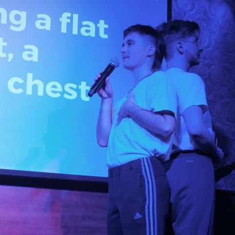 Two people standing back to back. One singing into a mic. Some words are readable on the back of the screen "a flat" and "chest".