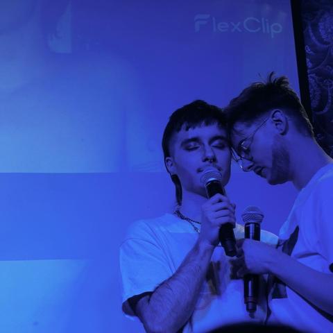 Two people on stage, one singing into the mic, the other leaning their head against the singers head.