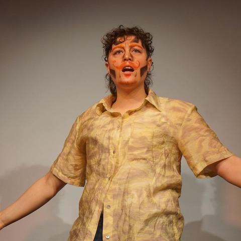 Person in yellow shirt standing on stage holding their arms wide open.