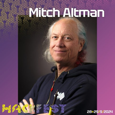 Portrait of hackerspace founder Mitch Altman smiling into the camera, his long white hair in partially dyed bright blue and red and holding a soldering iron