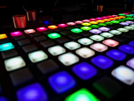 A close-up photo of the Synthstrom Deluge groovebox in a dark environment. Many of the colorful RGB-LED backlit pads are lit up. They reflect on the three knurled gold knobs on the device.