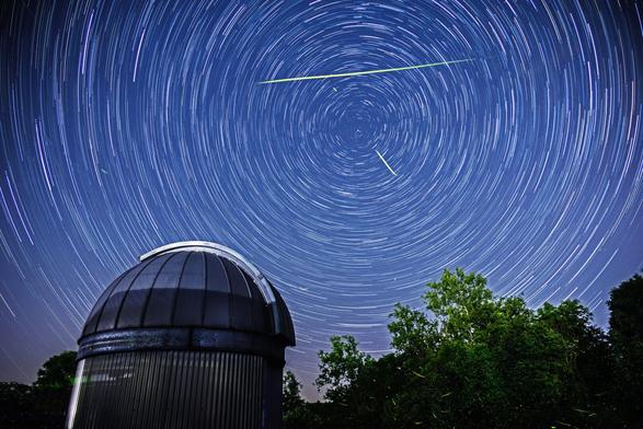 A photograph of star trails over a small observatory building. There are also lightning bug trails and one bright meteor.