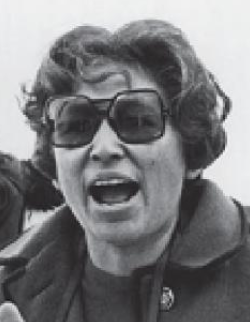 Tillie Walker at a protest in the 1960s; a woman with tan skin and dark hair in a set style, wearing sunglasses and a dark coat, in the process of speaking or shouting or singing