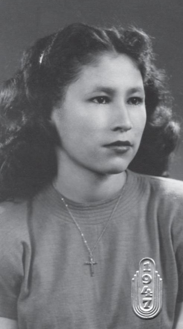 Tillie Fay Walker in her youth; a young Native American woman with wavy dark hair parted center and dressed loose but with combs to hold it away from her face; she is wearing a crew-neck short-sleeved sweater and a delicate gold cross on a chain