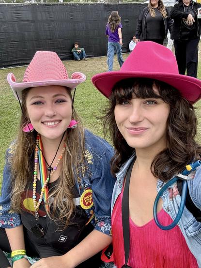 Selfie of two lesbian women in pink cowboy hats smiling at the camera sitting in the grass