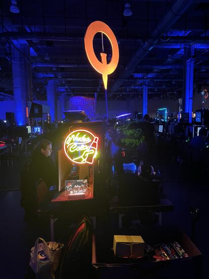 Photo of the Quakecon convention center room where the "bring your own computer" (BYOC) was held. It's a dimly lit room (lots of dark blues) with glowing neon signs, people seated at computers, and gaming equipment all along tables lining the hall. In the front is a neon sign that says "Nuka Cola" with a soda bottle.

A giant orange "Q" in the Quake style is hanging from the middle of the ceiling.