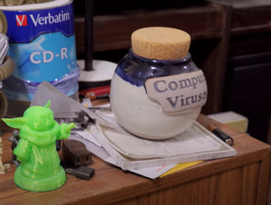 A table stacked with things. Notably, a large container of blank CD-roms, a green plastic Yoda figurine, and of most interest: A ceramic cookie jar labeled "Computer Viruses".