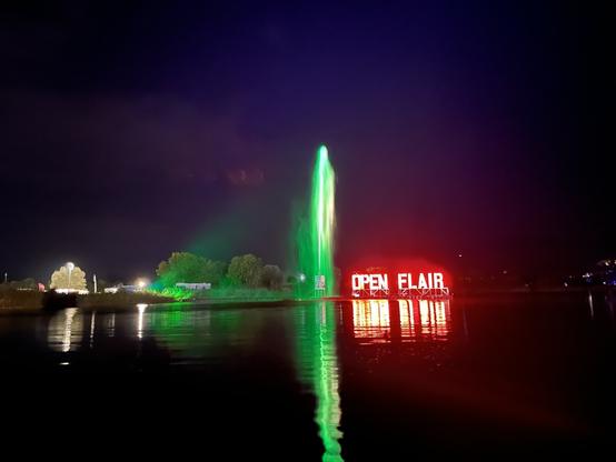 Ein Foto vom See neben der Seebühne beim Open Flair Festival bei Nacht. Eine Wasserfontäne, die grün angestrahlt wird, steigt neben einem rot leuchtenden Open Flair Schriftzug auf, der auf dem See schwimmt.