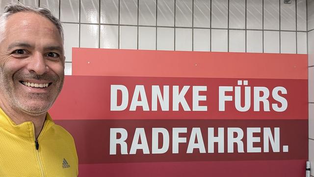 Ich im Rennrad-Trikot vor einer gefliesten wand mit einem Schild: Danke fürs Radfahren.