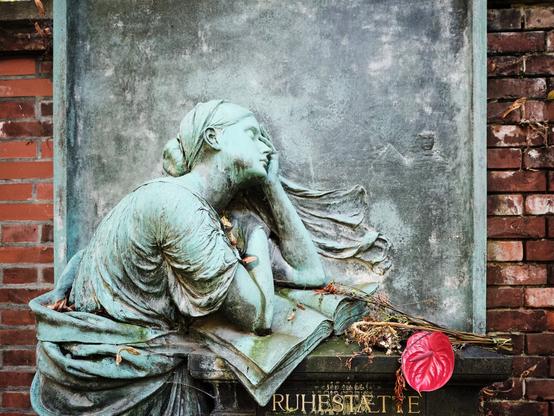 Das Bild zeigt eine bronzene Grabskulptur, die eine trauernde Frau darstellt. Die Figur lehnt nachdenklich auf einem offenen Buch, das vor ihr liegt. Ihr Kopf ruht in ihrer linken Hand, während ihr Blick in die Ferne gerichtet ist. Ihr Haar ist zu einem Knoten im Nacken gebunden, und ihr Gewand fällt in weichen Falten um ihren Körper.
Die Skulptur hat eine grünliche Patina, die typisch für gealterte Bronze ist. Vor der Statue liegen verwelkte Blumen, unter anderem eine rote Anthurie. Die Statue ist an einer Ziegelwand angebracht, und am unteren Rand ist das Wort „RUHESTÄTTE“ eingraviert, was in etwa „Ort der Ruhe“ bedeutet.
Insgesamt vermittelt die Szene eine Atmosphäre der Stille und des Gedenkens.