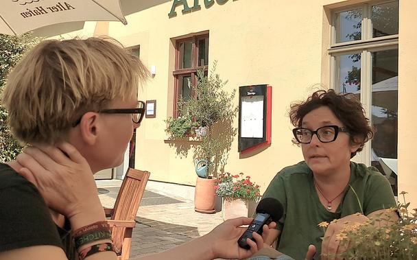 Eva Wolfangel von hinten mit einem Mikrofon in der Hand, dass sie Biella Coleman hinhält, an einem Tisch auf einer Terrasse. Über den beiden ein Sonnenschirm, auf dem klein und spiegelverkehrt steht "Alter Hafen".