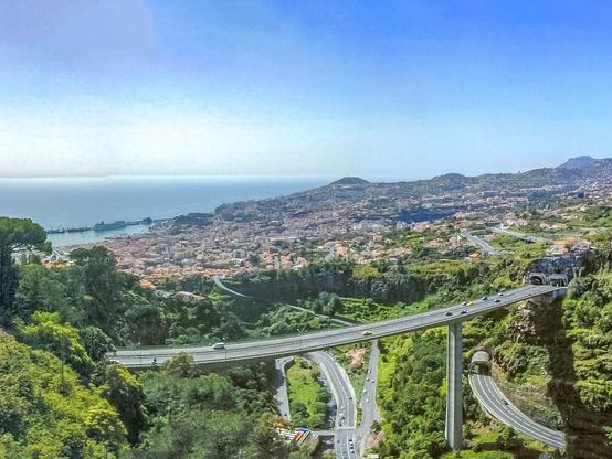 Das Bild zeigt den Blick herunter in die Bucht von Madeira mit Hafen. Im Vordergrund steht eine sehr hohe Autobahnbrücke, die über einer grün bewachsenen Schlucht zwei Tunnel miteinander verbindet. Darunter weitere breite Zufahrtsstrassen, die den Hang hinunter in die Stadt führen. Im Hintergrund erkennt man die dichtbebauten Stadtteile Funchals und links ein Stück Atlantik und der dunstblaue Himmel.
