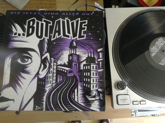 Rechts eine Platte auf dem Plattenspieler, links das Cover der LP "Bis jetzt ging alles gut" von ...But Alive: Ein Motiv von Eric Drooker zeigt ein halbes Gesicht vor Schienen mit einer Stadt im Hintergrund. Das Motiv hat den Stil eines Linolschnitts, Grundfarbe ist schwarz, die Stadt strahlt in weiss mit violette Crona.