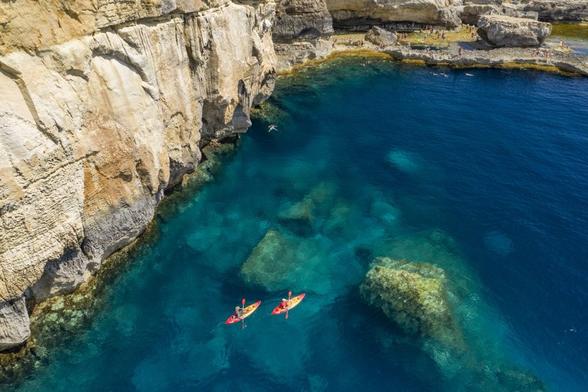 Malta, falesie a picco sul mare cristallino verde e blu sul quale remano due canoe