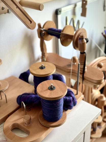 Two large wooden bobbins sitting on an upright lazy kate are mostly full with gorgeous purple-blue handspun yarn, flecked with teal and pink. A braid of the same fiber snakes between the bobbin toward the spinning wheel in the background with more of the same yarn on the bobbin-in-progress.
The lazy kate and the tall yarn swift behind sit on a set of white Ikea drawers below a weavibg warping board on the wall. And a whiteboard with large magnetic clips is blurred in the background.