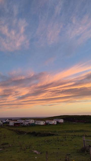 Blue sky with bars of cloud moving from left to right across the frame which are being backlit by a red sunset
There are some static caravans at the bottom of the picture and an expanse of grass in the foreground