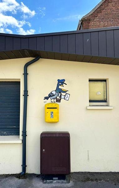 Streetart. A funny mural depicting a "carrier pigeon" in comic style was sprayed/painted on a yellow French letterbox on a lower building. It is a simply drawn pigeon with a rather unusual blue head. It has a protruding lower jaw, a large white eye and a small black eye. It is wearing a blue letter carrier's cap and a corresponding bag. She is holding a letter in her hand with one wing. Hopefully she will find the recipient.
Info: The anonymous artist "TocToc" is a French freelance illustrator and video artist. He exhibits his work in galleries around the world. In 2012, he created the simple figures DUDUSS when he was an art student and still sprays it on the walls of cities today.