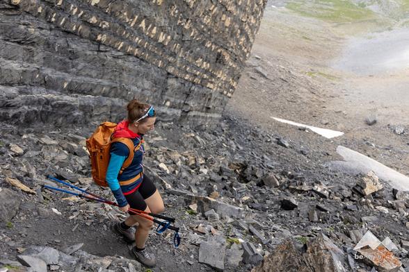 Woman in hiking clothes and an orange backpack walking down a scree slope. View of the scene from above. Rock wall on the left in the picture.