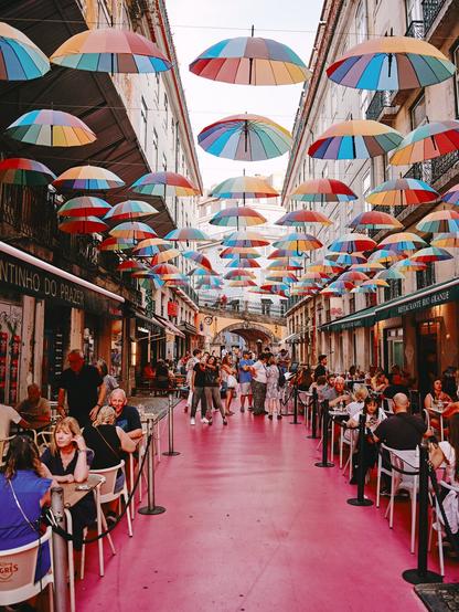Die berühmte Pink Street in Lissabon, Portugal, erstrahlt in einer lebendigen Atmosphäre. Über den Köpfen der Besucher hängen bunte Regenschirme, die das urbane Flair dieser beliebten Straße unterstreichen. Die Mischung aus kreativer Gestaltung und pulsierendem Stadtleben macht diesen Ort zu einem Highlight für Einheimische und Touristen gleichermaßen.