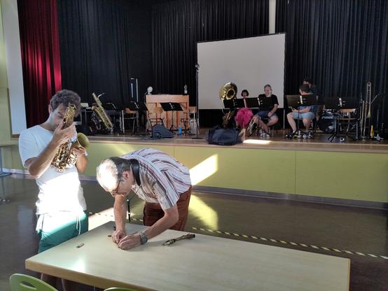 Zwei Saxophonisten hantieren mit Werkzeug an einem Altsax herum.
Im Hintergrund wartet auf der Bühne der schwitzende, aber trotzdem außergewöhnlich gutaussehende Rest des Orchesters darauf, daß es endlich weitergeht