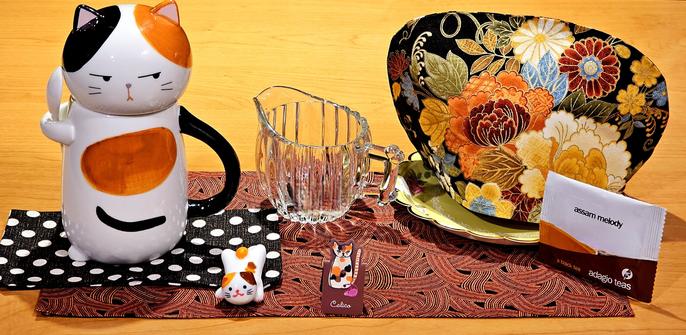 A tea tableau devoted to calico cats. The mug is a figural cat in calico colors of white, black, orange, and brown. There is a small china figurine of a calico cat, and a tiny painting of one. The tea is Assam Melody from Adagio