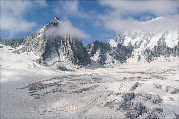 Huge pyramidal mountain emerges from a crevasses glacier. Tow mountaineers make their way from left to right seeming tiny against the scale of the mountains.