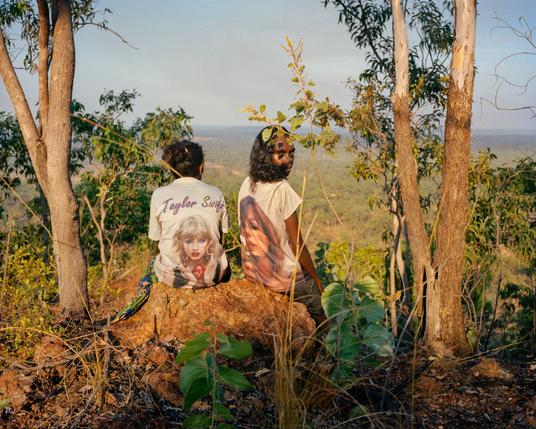 Cousin sisters Shauna and Bridget Perdjert from Kardu Thithay Diminin Clan and Murrinhpatha language group, sit on Air Force Hill after practicing cultural burning on Kardu Yek Diminin Country, Wadeye, Northern Territory. 

Both girls, wearing Taylor Swift T shirts, are sitting on a hilltop boulder between two trees, looking out over green scrubby country below.