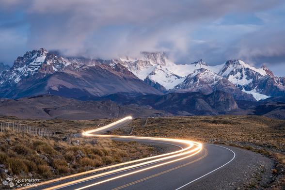 S-curved road through grassland vanishing over a hill. The white light trail of a car follows that road. In the background, one can see a mountain range with the tops hidden in clouds.