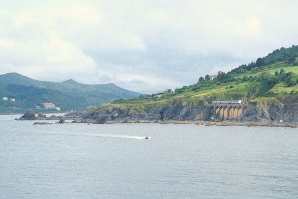 coastline with lot of sea. Tiny in the background an Euskotren service is visible on a railway viaduct. Green hills further in the background. Cloudy sky.