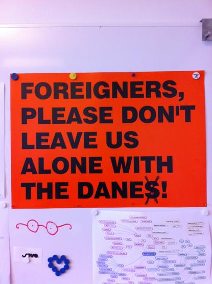 A large red sign, with bold, black lettering, attached to a white board with magnets. It reads "Foreigners, please don't leave us alone with the danes" the S of danes has been crossed out. Other things below it on the white board include a badly drawn pair of spectacles and a a group of magnets that have been made into a heart.