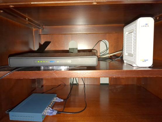 A photograph of the inside of a TV cabinet. On the top shelf we have a MikroTik hAP ac3 router next to an ARRIS SURFboard modem. On the bottom shelf we have a TP-Link ethernet switch.
