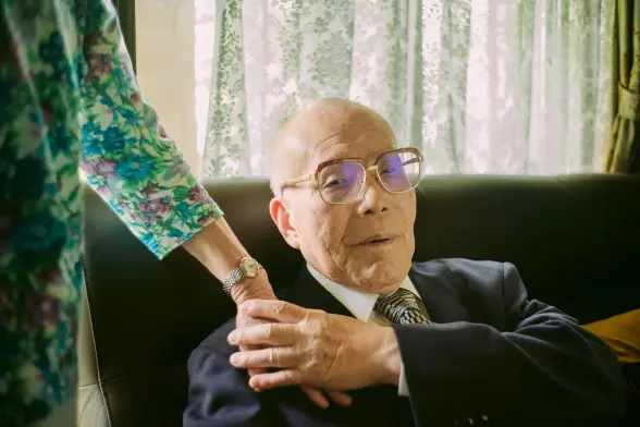 A poignant portrait of a survivor of the Hiroshima bombing in Japan. He is seated in the couch wearing a suit. He is looking at the camera while holding his wife's hand on his shoulders. Photograph by Kentaro Takahashi