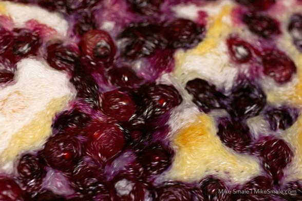 A blueberry cobbler with stylistic swirls added in post production as it cools on a stove top. Golden islands of sugary topped pastry break up the sea of purple and red berries.
