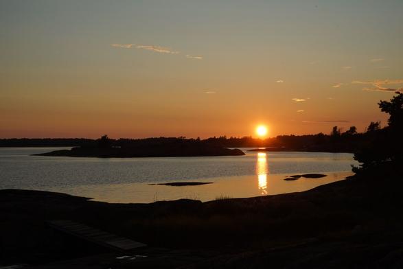 Photo of a sunset over a small bay. In the foreground and at the horizon the landscape is almost black, the sun is about to hide behind trees in the distance. The reflection of the sun in the water looks almost like a straight line but a bit of wind makes the surface of the water a bit curly in the middle.