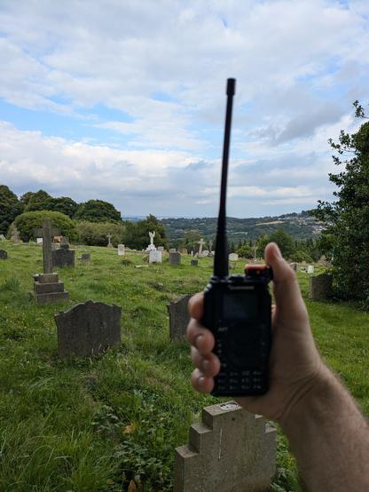 A hand holding a handheld transceiver up to the sky in a cemetery with headstones, green grass and a partially cloudy blue sky in the background