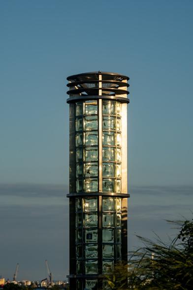 A color photograph of a glass and steel lightning column on a pedestrian walkway over an interstate highway. The cylinder is mostly composed of glass blocks within a thin steel structure. The blue sky and thin clouds behind are getting lightly darker, and the late afternoon sunlight is hitting the glass from the right. At the bottom in the background, cranes and storage tanks from the port are visible, along with just a hint of blue water from the Providence River.