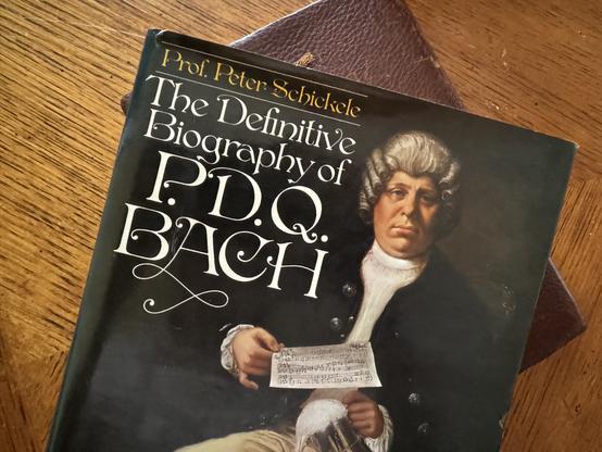The 1976 hardcover edition of “The definitive biography of PDQ Bach” by Peter Schickele. It shows a painting of a man in baroque attire and wig holding a leaf of sheet music.