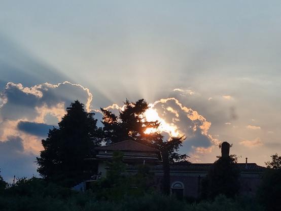 Tramonto dietro le nuvole, dietro gli alberi 🌲 dietro una 🏡, mentre la luce irraggia il panorama