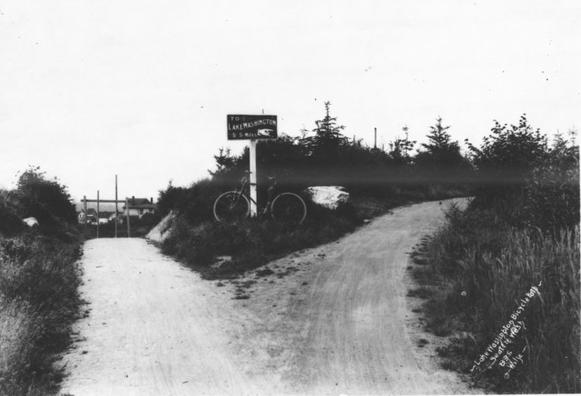 Old black and white photo of a fork in a bike path with a sign pointing right that reads To Lake Washington 5.5 miles.