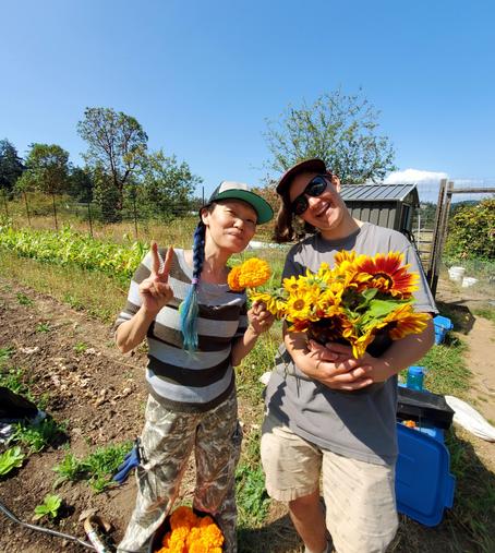 Photo of me & Ardeo, queer farm owner, in farm fields. I'm holding 2 marigold flowers & Ardeo is holding a bucket of cut sunflowers.