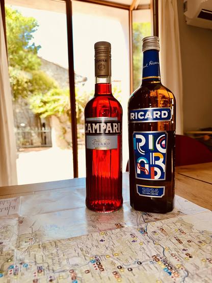 A bottle of Campari and a bottle of pastis (Ricard) on a table in front of a sunny window.
