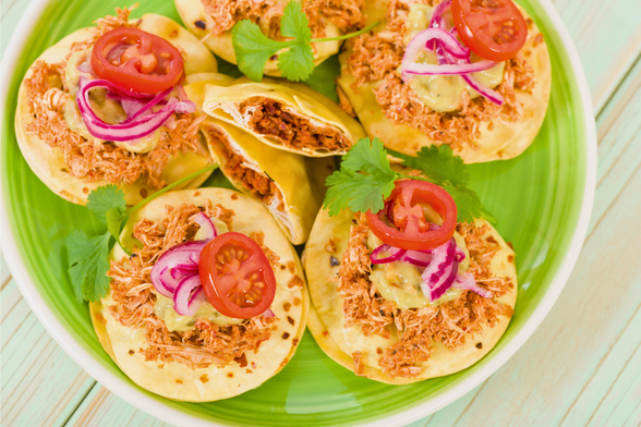 A plate with Yucatan panuchos topped with shredded chicken, guacamole, pickled red onion and tomato. One of them is halved, showing the bean filling.