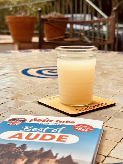 A glass of pastis on a tiled table outdoors. Next to it is a guidebook to the Aude department that was given out for free at Carcassonne airport.
